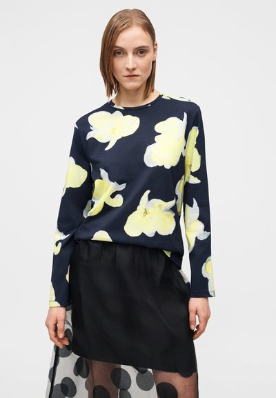Σκούρο μπλε μπλούζα με μακριά μανίκια και μεγάλο κίτρινο λουλουδένιο σχέδιο· μαλακό υλικό· φοριέται πάνω από μαύρη διαφανή φούστα με πουά σχέδιο.