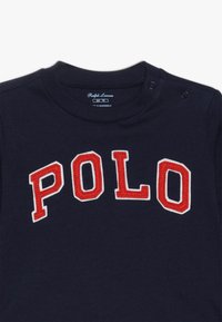 Dětská mikina z bavlny v námořnické modré barvě s žebrovaným výstřihem, na přední straně s vyšitou aplikací "POLO" v tučném červeném a bílém písmu. Dva patentky na rameni.