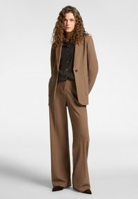 Donna con capelli ricci che indossa pantaloni larghi marroni e un blazer abbinato su una camicia nera con bottoni, in piedi con le mani dietro la schiena.