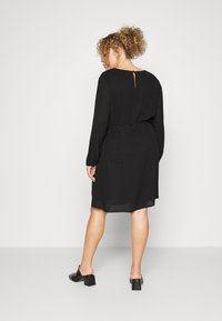 Vestido preto de mangas longas com cintura cintada, recorte em forma de chave redonda nas costas e barra fluida. Usado com sapatos pretos de salto bloco.