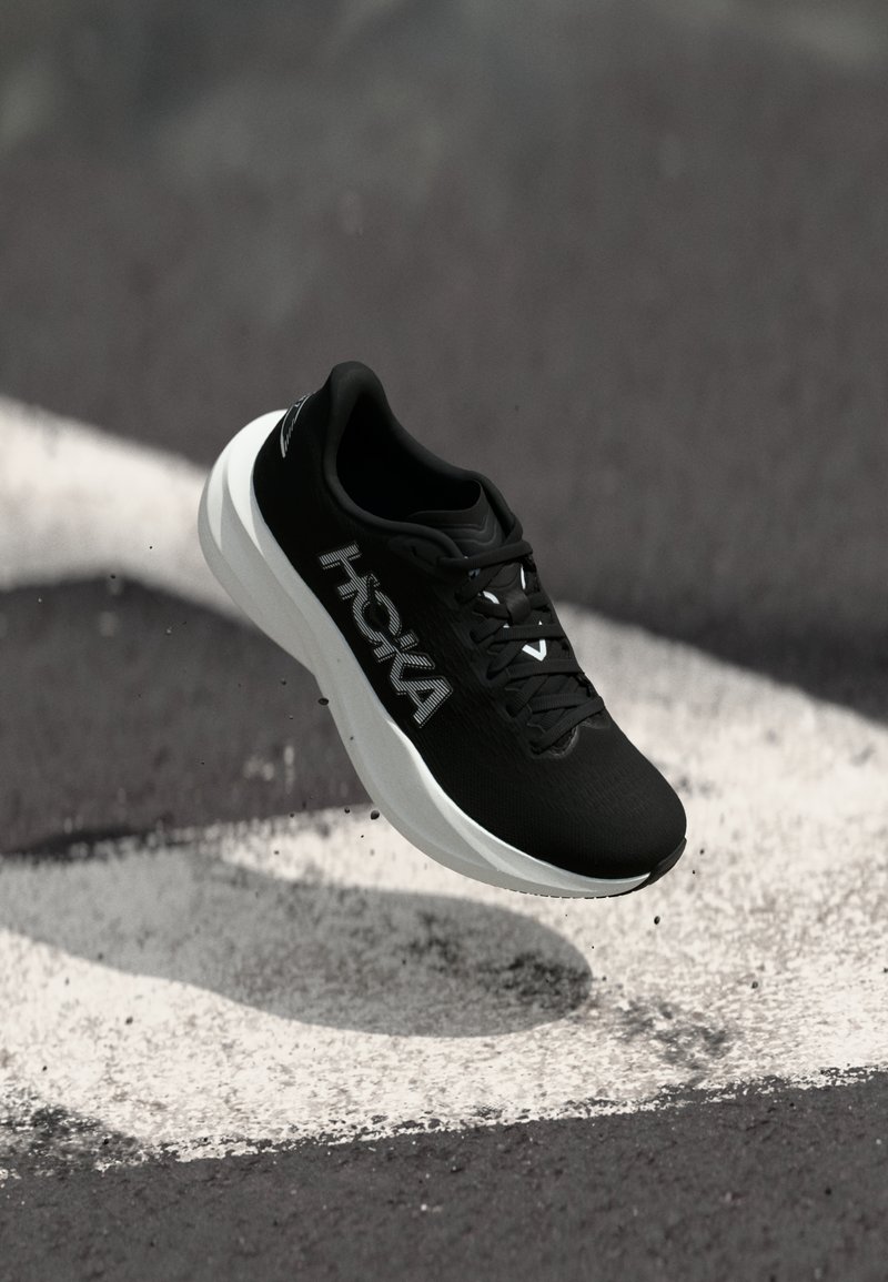 Μαύρο αθλητικό παπούτσι Hoka με λευκή σόλα στον αέρα πάνω από άσφαλτο με λευκή γραμμή.