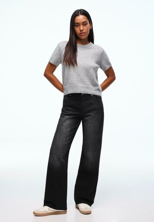 Szary sweter z krótkim rękawem o miękkiej fakturze, zestawiony z czarnymi szerokimi dżinsami. Modelka ma na sobie białe trampki z beżowymi akcentami.