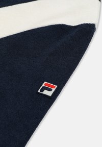 Marineblaues und weißes Handtuckseck mit einem kleinen, gestickten FILA-Logo-Patch in Rot, Marineblau und Weiß auf texturierter Stoffoberfläche.
