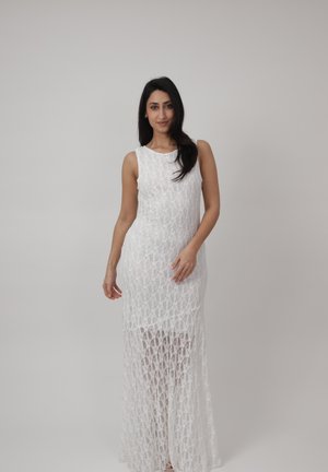 Frau trägt ein ärmelloses weißes Maxikleid aus Spitze, steht mit einer Hand seitlich und berührt mit der anderen das Kleid, neutraler Hintergrund.