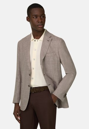 Mand iført brun ternet blazer, cremefarvet poloshirt med knapper, brun bælte og brune bukser, ser til siden med den ene hånd i lommen.