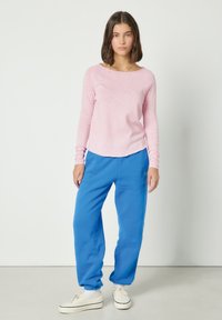 Mladá žena stojící, oblečená v světle růžovém tričku s dlouhým rukávem, jasně modrých kalhotách, bílých ponožkách a bílých teniskách na jednobarevném pozadí.