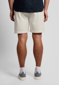 Οπίσθια όψη ατόμου που φορεί μπεζ σορτς, σκουρόχρωμο πουκάμισο, λευκές κάλτσες και μπλε πάνινα παπούτσια, μπροστά σε απλή φόντο.