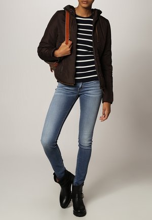 Femme portant une doudoune noire, un chemisier rayé noir et blanc, un jean bleu clair, des bottines noires et un sac à bandoulière marron.