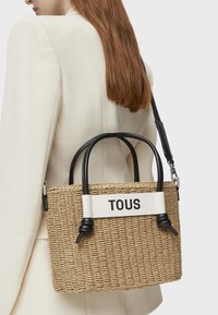 Woven straw handbag with black leather handles and strap. Features a white band with the text "TOUS." Compact rectangular shape.