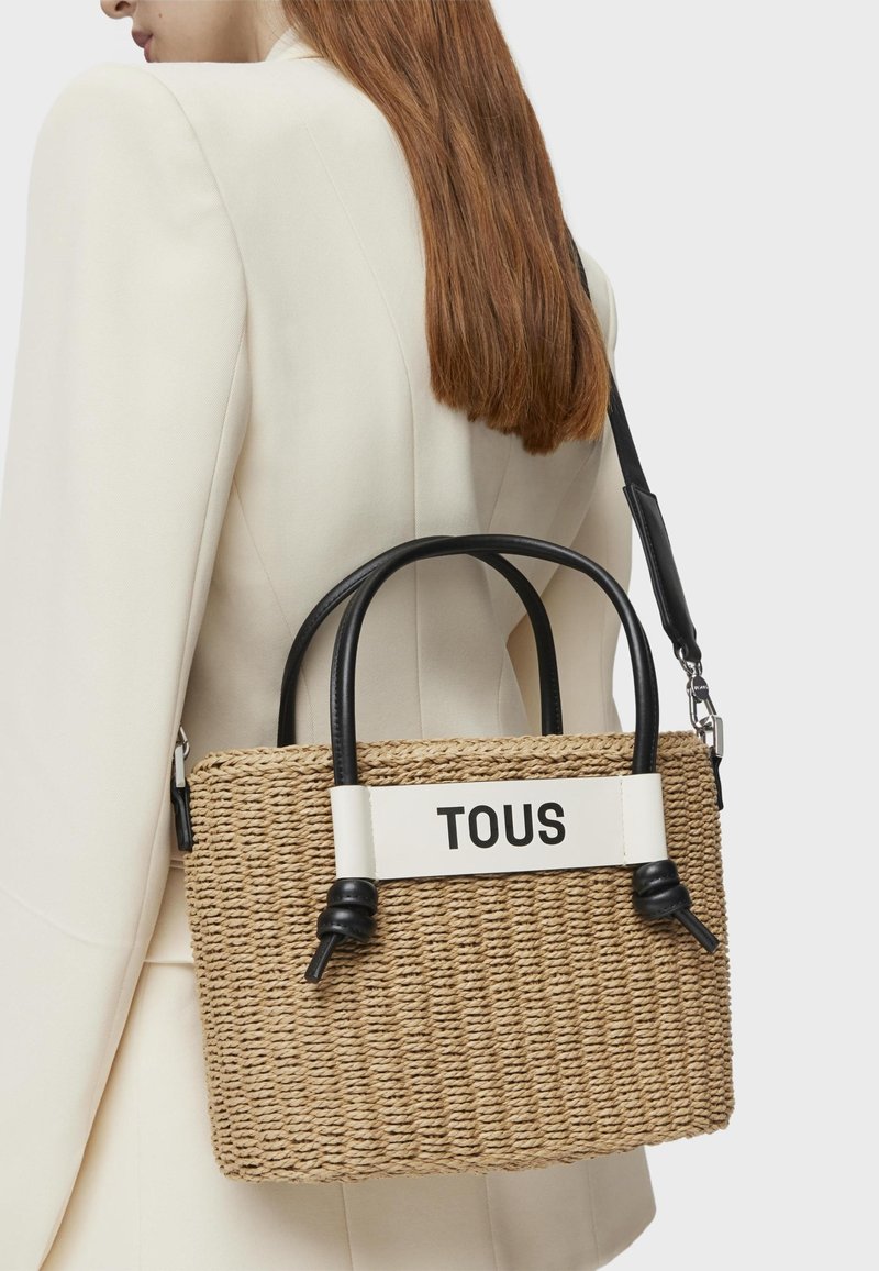 Woven straw handbag with black leather handles and strap. Features a white band with the text "TOUS." Compact rectangular shape.
