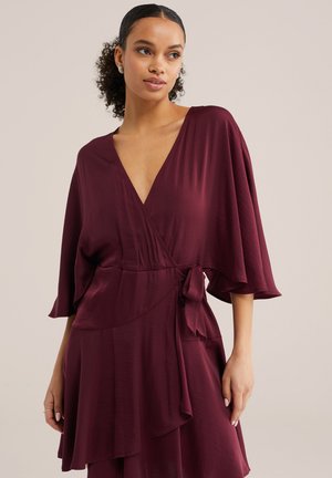 Femeie cu păr creț, purtând o rochie maro în încheietură cu decolteu adânc în V, mâneci largi și o fundă legată pe lateral, stând în fața unui fundal simplu.