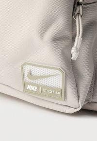 Γωνία τσάντας Nike Utility 2.0 σε μπεζ με υφασμάτινη υφή, εμφανές φερμουάρ και ορθογώνιο έμβλημα με το λογότυπο Nike και το swoosh.