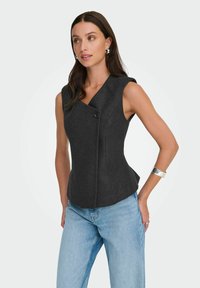 Top senza maniche grigio scuro su misura con scollatura a V e chiusura con un bottone; tessuto liscio con cuciture laterali, abbinato a jeans azzurri.