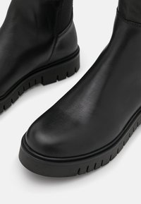 Botas de tobillo de cuero negro con un acabado liso, punta redonda y una suela gruesa y texturizada que presenta surcos profundos para mayor tracción.