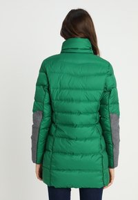 Grön puffjacka med hög krage, med gråtygsdetaljer på ärmarna. Designen är vadderad med synliga horisontella sömmar.