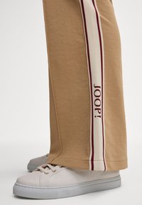 Khakifarbene Trainingshosen mit einem weißen Seitenstreifen, der mit maronenfarbener Schrift "JOOP!" bedruckt ist, aus glattem Baumwollmaterial, kombiniert mit weißen Sneakers.