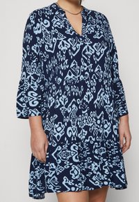 Mörkblå klänning med ett ljusblått abstrakt mönster. Har V-urringning, trekvartsärmar och en utsvängd nederkant med draperad midja.