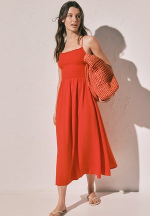 Kobieta w czerwonej sukience midi bez rękawów, trzymająca teksturowany czerwony sweter, stoi przy jasnej ścianie, za nią pada cień.