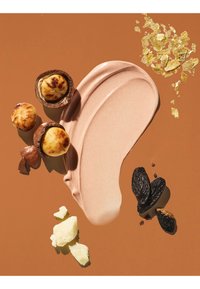 Tache de crème cosmétique beige crémeuse entourée de noix de macadamia, flocons d’or, morceaux de beurre de karité et fèves tonka noires sur fond marron.