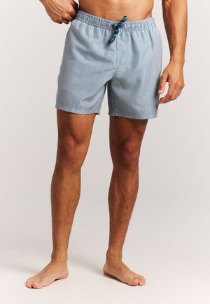 Homme portant un short à cordon de serrage bleu clair et blanc à fines rayures, debout pieds nus sur un fond uni.