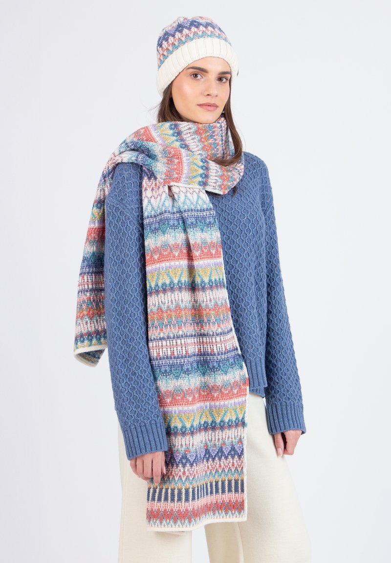 Gehaakte muts en sjaal set in het blauw, met gekleurde geometrische patronen. Gezellige textuur met geribbelde randen. Model draagt het met een blauwe trui.