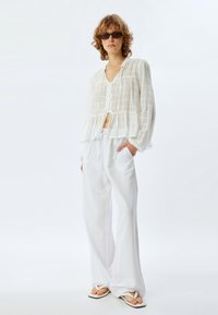 Witte blouse met knopen, voorzien van een gelaagd ontwerp, lange mouwen en lichte stof. Gepaarde met witte broek met trekkoord en slippers.