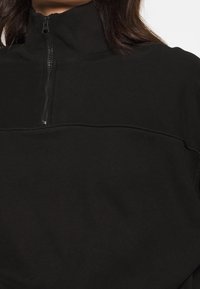 Sweat-shirt noir à fermeture éclair avec un col montant, doté d'un tissu texturé et d'une couture horizontale traversant le devant. Design minimaliste.