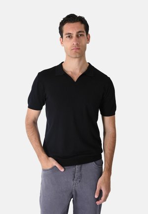 Uomo in piedi di fronte, indossa una polo nera a maniche corte e pantaloni grigi con una mano in tasca su sfondo bianco.