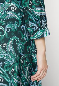 Main à peau pâle reposant à côté d'un vêtement vert foncé à motif cachemire avec des détails bleus et blancs et un revers retroussé.