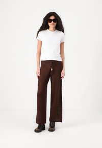 Bílé tričko, hnědé pletené kalhoty s tkaničkou a dřevěnými akcenty. Stylizováno s tmavými oversized slunečními brýlemi a tmavými kotníkovými botami.