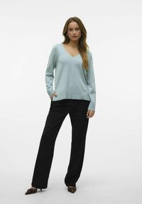 Maglione azzurro chiaro con scollo a V e maniche lunghe, abbinato a pantaloni neri a gamba larga e scarpe con il tacco a punta e dettagli decorativi.