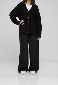 Cárdigan negro de punto con un patrón de cable texturizado, escote en V, con seis botones en el frente, combinado con pantalones holgados negros y zapatillas blancas.