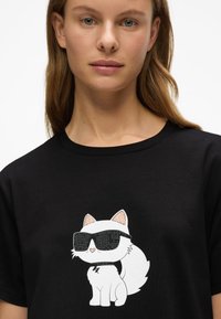 Γυναίκα που φοράει μαύρο μπλουζάκι με γραφικό λευκού καρτούν γάτας, φορώντας μαύρα γυαλιά ηλίου με καρφιά και κολάρο.