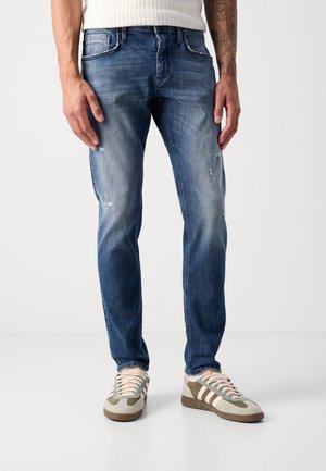 Hombre vistiendo jeans ajustados de mezclilla azul deslavada con un ligero desgaste y zapatillas beige con franjas blancas y verde oliva.