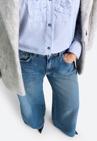 Szerokie niebieskie dżinsy z wyblakłym teksturą, połączone z niebieską i białą w paski koszulą oraz szarym wełnianym żakietem.