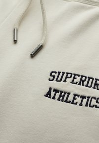 Crème-kleurige sweatshirt met "SUPERDRY ATHLETICS" geborduurd in donkerblauw, voorzien van metalen uiteinden aan de koorden en een zachte textuur.
