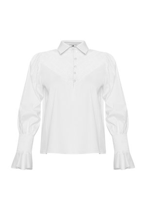 Blusa blanca de manga larga con cuello tipo camisa, tapeta de botones, yugo texturizado, mangas abullonadas y puños con volantes. Tejido de algodón suave.