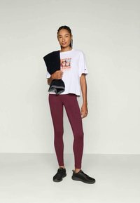 White graphic t-shirt with a retro design, paired with fitted burgundy leggings and black athletic shoes. A dark towel is draped over one shoulder.