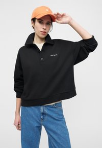 Άτομο που φοράει πορτοκαλί καπέλο, μαύρο πουλόβερ Carhartt με γιακά και μπλε τζιν, ποζάροντας με το ένα χέρι να ακουμπάει το καπέλο.