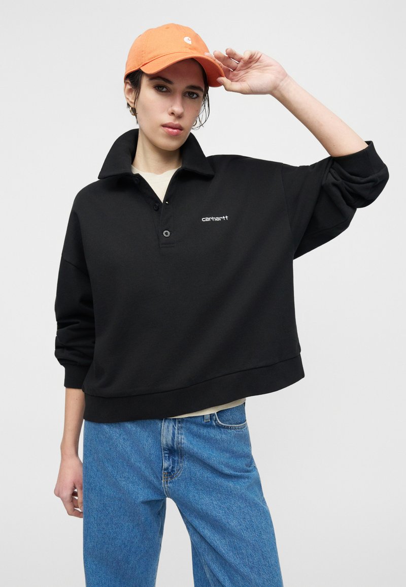 Personne portant une casquette orange, un pull Carhartt noir avec col et un jean bleu, posant avec une main touchant la casquette.