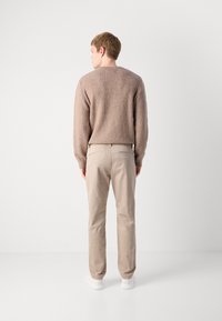 Mann, der mit dem Rücken zur Kamera steht, trägt einen strukturierten braunen Pullover, beigefarbene Hosen und weiße Turnschuhe vor einem schlichten weißen Hintergrund.
