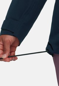 Navy jacket sleeve with adjustable cuff; hand holding a dark green drawstring. The material appears smooth and lightweight.