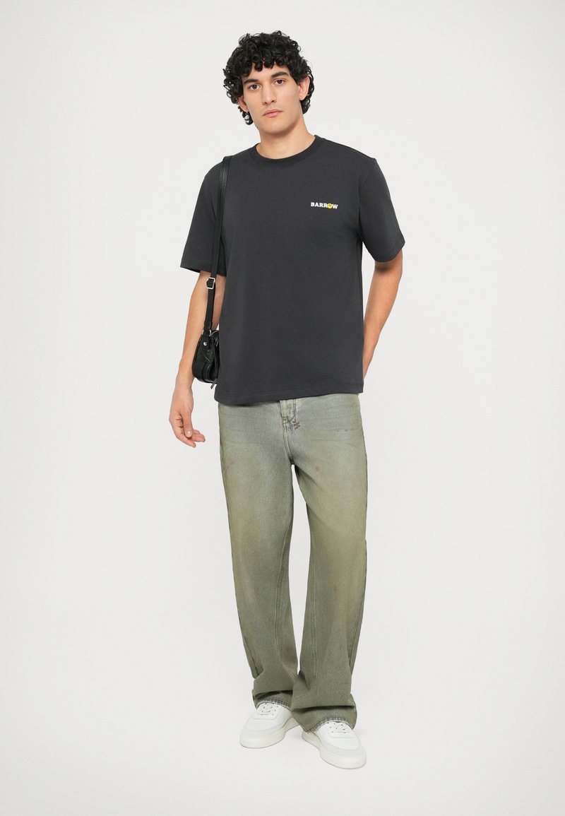 T-shirt nera in cotone con logo "BARROW" multicolore sul petto, abbinata a jeans a gamba larga verde chiaro e sneaker bianche.