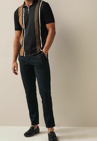 Polo noir à rayures verticales marron et blanches, manches courtes, col zippé. Associé à un pantalon foncé et des mocassins noirs.