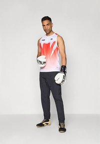 Uomo che indossa una maglietta sportiva senza maniche bianca e rossa, pantaloni neri, guantoni da boxe e scarpe sportive nere, in piedi su uno sfondo semplice.