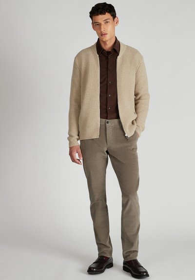 Man in beige ritsbrei cardigan, donkerbruine button-up shirt, lichtbruine corduroy broek en donkerbruine loafers, staande tegen een effen achtergrond.