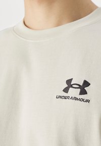 Ljust beige sweatshirt med rund halsringning, som har en svart broderad Under Armour-logga på bröstet. Mjuk, strukturerad tyg.
