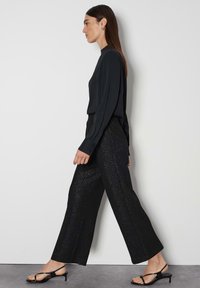Zwarte top met lange mouwen gecombineerd met zwarte broek met een sprankelende textuur. Model draagt zwarte sandaaltjes met bandjes. Effen achtergrond.