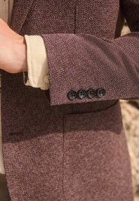 Manteau à manches texturé marron présentant quatre boutons noirs, porté sur une chemise de couleur claire. Détails de tissage remarquables dans le design du tissu.