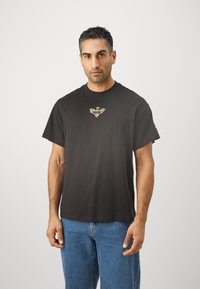 Sort bomulds t-shirt med rund hals og korte ærmer, med et centralt guld emblemdesign. Sammen med lyseblå denim jeans.
